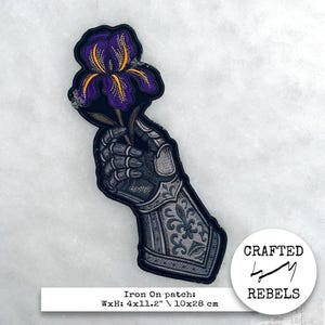 May include: Iron-on patch featuring a detailed illustration of a metallic gauntlet holding a purple iris flower with yellow accents. The patch is 4 x 11.2 inches (10x28 cm) and includes the text "Crafted Rebels".