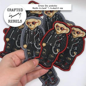May include: A collection of embroidered iron-on patches featuring a tan teddy bear wearing red sunglasses, a black leather-style jacket, and distressed black jeans with a silver chain. Each patch measures 5.4 by 10 cm, labelled Crafted Rebels.