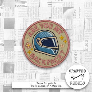 Może przedstawiać: Haftowana naszywka z napisem "ARE YOU MY BACKPACK?" z niebieskim wzorem kasku, różową obwódką i małymi akcentami kwiatowymi i gwiazdkami. Mierzy 9x9 cm.