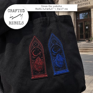 May include: Two embroidered iron-on patches attached to a black fabric tote bag. Each patch features a pointed gothic arch design with a crescent moon and mountain landscape. One patch is red, the other is blue. Dimensions are 6x17 cm.