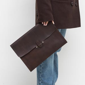 Könnte beinhalten: Ein dunkelbrauner, zweireihiger Blazer und eine passende Ledermappe. Die Person trägt eine blaue Jeans und beige Ballerinas. Der Blazer hat zwei Knopfreihen und ein fallendes Revers. Die Mappe hat einen Klappenverschluss.