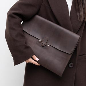 May include: A dark brown leather portfolio with a flap closure and metal hardware. The portfolio is held in front of a person wearing a brown blazer and blue jeans. The background is a plain white wall.