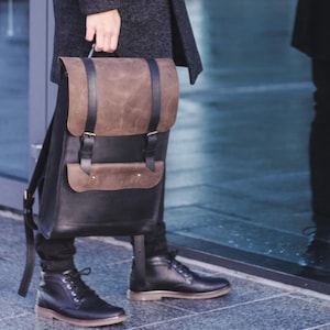 Puede incluir: Un abrigo gris oscuro y una mochila negra con detalles de cuero marrón. La mochila tiene un cierre de solapa y correas ajustables. La persona lleva botas negras y está de pie junto a una pared de cristal.