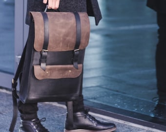 May include: A dark gray coat and black backpack with brown leather accents. The backpack has a flap closure and adjustable straps. The person is wearing black boots and standing outside near a glass wall.