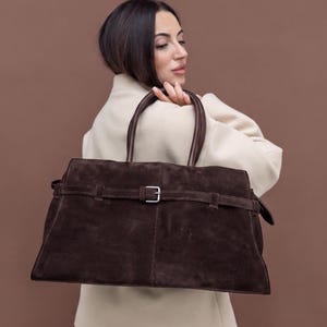 May include: A large dark brown suede handbag featuring two rounded top handles and a horizontal strap across the front secured with a silver-toned metal buckle. The bag has a rectangular shape with visible stitching details along the center seam.