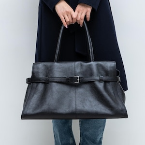 May include: A black leather tote bag with a buckle fastening and top handles. The bag has a textured surface and a rectangular shape. The person is wearing blue jeans and a navy coat.