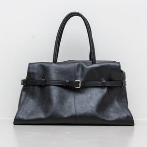 Könnte beinhalten: Eine schwarze Ledertasche mit einem oberen Griff und einem Schnallenverschluss. Die Tasche hat eine rechteckige Form mit einer strukturierten Oberfläche. Die Tasche hat einen Riemen, der um die Mitte der Tasche mit einer silbernen Schnalle verläuft.
