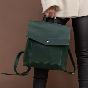 Puede incluir: Una mochila de cuero verde oscuro con cierre de solapa y botón dorado. La mochila tiene correas ajustables y un asa superior. La persona lleva pantalones negros, una chaqueta color crema y botines de tacón color beige.