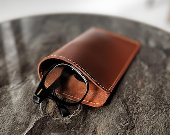Puede incluir: Un estuche de gafas de cuero marrón con costuras blancas, que contiene unas gafas con montura negra. El estuche es rectangular y descansa sobre una superficie gris texturizada. Las gafas son parcialmente visibles.