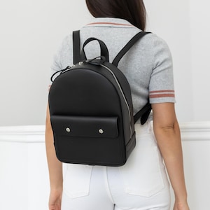 Könnte beinhalten: Ein schwarzer Rucksack mit abgerundeter Oberseite und einer Vordertasche mit zwei silbernen Knopfverschlüssen. Der Rucksack hat einen silbernen Reißverschluss und schwarze Schultergurte. Die Person trägt ein graues Hemd und eine weiße Hose.