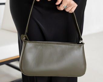 May include: Olive green shoulder bag with a rectangular shape and a long, thin strap. The bag is held by a person wearing a black hoodie and black pants. The bag has a smooth, matte finish.