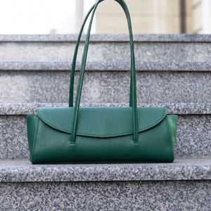 May include: A green leather handbag with a flap closure and two long handles. The bag is rectangular with a textured surface and is on grey stone steps.