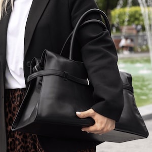 Op de afbeelding: Een gestructureerde zwarte leren handtas met twee handvatten, een horizontale riem met metalen gesp en een hoofdvak met ritssluiting. De tas heeft een gladde afwerking en een rechthoekige basis in een strak en eenvoudig ontwerp.