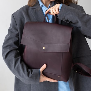 Peut inclure: Un sac à dos en cuir bordeaux avec une fermeture à rabat et un bouton doré. Le sac à dos a une poignée supérieure et des bretelles réglables. Le sac est tenu par une personne portant un blazer gris et une chemise bleue.