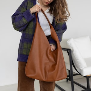 Könnte beinhalten: Eine große, braune Ledertasche mit Schulterriemen. Die Tasche wird von einer Person gehalten, die ein weißes Hemd, eine braune Hose und eine blau-grüne karierte Strickjacke trägt. Die Tasche hat ein schlichtes, minimalistisches Design.