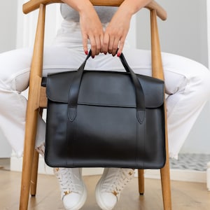 May include: A black leather briefcase with a top handle and a flap closure. The briefcase is held by a person sitting on a wooden chair. The person is wearing a gray polo shirt, white pants, and white sneakers.