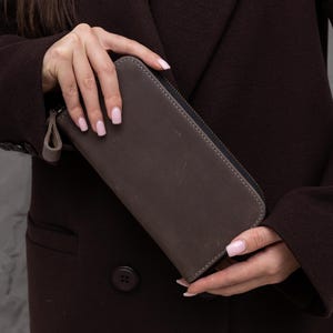 Könnte beinhalten: Eine braune Ledergeldbörse mit Reißverschluss. Die rechteckige Geldbörse scheint aus einem Leder mit Used-Look zu sein. Der Hintergrund ist ein dunkelbrauner Mantel. Die Geldbörse wird von einer Person gehalten.