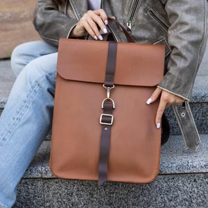 Può includere: Uno zaino in pelle marrone con chiusura a patta e cinturino verticale fissato da una fibbia color oro. Lo zaino ha un design minimalista e una forma rettangolare. La borsa è realizzata in pelle liscia.