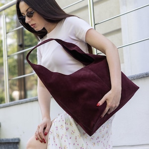 May include: A large, deep burgundy hobo-style shoulder bag crafted from a soft, matte suede material. The bag features a wide, integrated shoulder strap and a spacious, unstructured silhouette with a clean, minimalist design and no visible hardware.