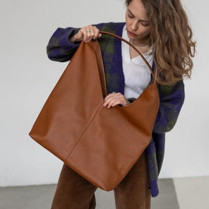 Può includere: Una grande borsa tote in pelle marrone con manico curvo. La borsa ha un design minimalista e un interno spazioso, adatta all'uso quotidiano. La pelle ha un aspetto strutturato.