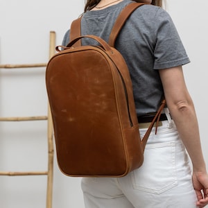 May include: A brown leather backpack with a rounded top and a vertical zip. The backpack has adjustable shoulder straps and a top handle. The leather has a distressed appearance. The person in the image is wearing a grey t-shirt and white jeans.