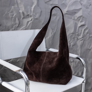 Peut inclure: Un sac à bandoulière en daim marron foncé avec une anse incurvée. Le sac est posé sur une chaise blanche avec une structure argentée. Le fond est un mur gris texturé.
