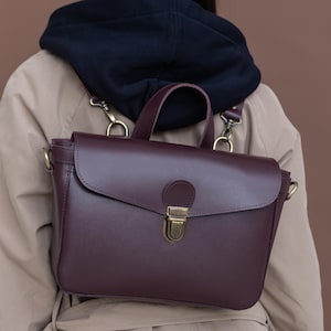 Pode incluir: Mochila de couro bordô estilo pasta com alça superior e alças ajustáveis. A bolsa possui fecho de aba com fecho de latão e ferragens de latão. A bolsa é usada sobre um trench coat bege e um moletom com capuz azul marinho.