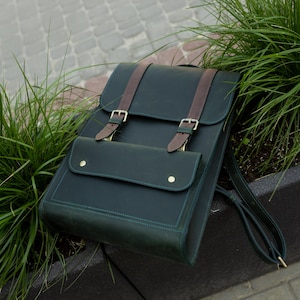 Puede incluir: Una mochila de cuero verde oscuro con solapa rectangular, dos correas de cuero marrón con hebillas metálicas plateadas y un bolsillo frontal asegurado con dos botones a presión de color latón. Incluye correas de hombro a juego.