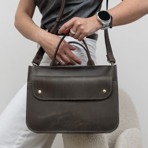 Può includere: Una borsa a tracolla in pelle marrone scuro con una tasca con patta e due borchie color ottone. La borsa ha un manico superiore e una lunga tracolla regolabile. La borsa è tenuta da una persona che indossa una maglietta grigia e pantaloni bianchi.