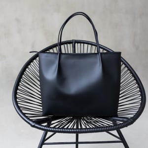 May include: A black tote bag with two handles rests on a black, woven chair. The bag is rectangular with a smooth, matte finish. The chair has a rounded back and a webbed seat, creating a contrasting texture.
