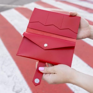 May include: A vibrant red leather wallet, open to show card slots and a snap closure. The wallet is held against a background of red and white stripes.