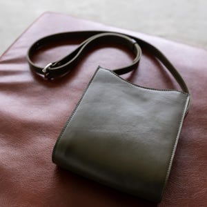 May include: A dark olive green leather crossbody bag with a long, adjustable strap. The bag has a rectangular shape with a curved top edge and a silver-colored buckle. The bag is sitting on a reddish-brown surface.