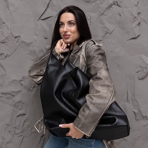 Puede incluir: Un bolso hobo negro grande con textura granulada, que presenta una silueta holgada, una correa ancha para el hombro y un compartimento principal espacioso. El bolso est&aacute; fabricado con un material flexible que se pliega de forma natural al sostenerlo.
