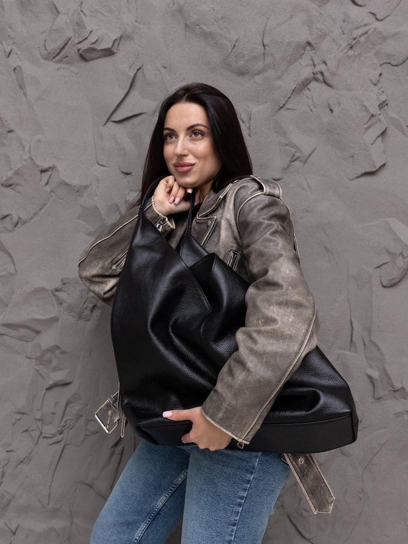 Puede incluir: Un bolso hobo negro grande con textura granulada, que presenta una silueta holgada, una correa ancha para el hombro y un compartimento principal espacioso. El bolso est&aacute; fabricado con un material flexible que se pliega de forma natural al sostenerlo.