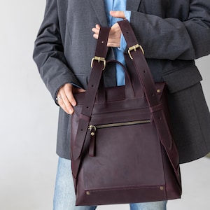 May include: A burgundy leather backpack with adjustable straps and gold-colored hardware. The bag features a zippered front pocket and a top handle. The design is minimalist and modern, suitable for everyday use.