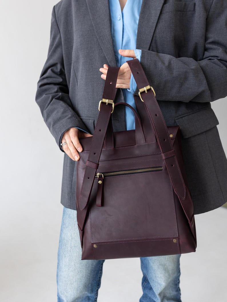 May include: A burgundy leather backpack with adjustable straps and gold-colored hardware. The bag features a zippered front pocket and a top handle. The design is minimalist and modern, suitable for everyday use.