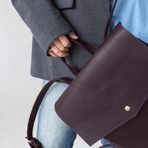 May include: A deep burgundy leather satchel with a flap closure and a gold-colored button. The bag has a shoulder strap and a top handle. The person is wearing a gray blazer, blue shirt, and jeans.