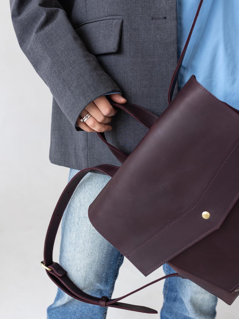 May include: A deep burgundy leather satchel with a flap closure and a gold-colored button. The bag has a shoulder strap and a top handle. The person is wearing a gray blazer, blue shirt, and jeans.