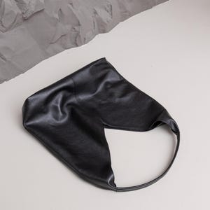 K&ouml;nnte beinhalten: Eine schwarze Leder-Umh&auml;ngetasche mit einem einzigartigen, asymmetrischen Design. Die Tasche hat einen einzelnen, abgerundeten Riemen und eine strukturierte Oberfl&auml;che. Die Tasche liegt auf einer wei&szlig;en Oberfl&auml;che mit grauem, strukturiertem Hintergrund.