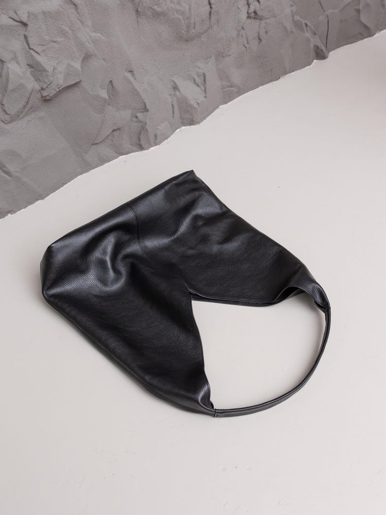 K&ouml;nnte beinhalten: Eine schwarze Leder-Umh&auml;ngetasche mit einem einzigartigen, asymmetrischen Design. Die Tasche hat einen einzelnen, abgerundeten Riemen und eine strukturierte Oberfl&auml;che. Die Tasche liegt auf einer wei&szlig;en Oberfl&auml;che mit grauem, strukturiertem Hintergrund.