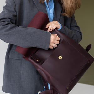 May include: A burgundy leather backpack with a flap closure and gold-colored hardware. The interior is a contrasting burgundy color. The backpack has adjustable straps and a top handle. The person is wearing a gray blazer, a light blue shirt, and blue jeans.
