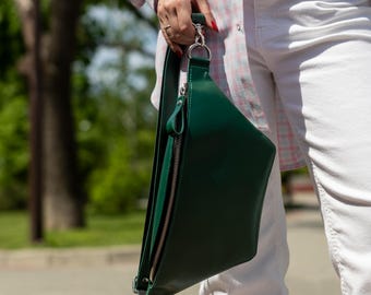 May include: A dark green crossbody bag with a smooth finish, featuring a silver-toned metal zipper closure, a matching green strap, and a silver swivel clasp attachment. The bag has a structured, triangular silhouette and visible stitching.