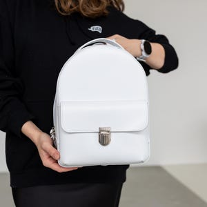 Puede incluir: Una mochila blanca con cierre de solapa y un cierre plateado. La mochila tiene un asa superior y correas ajustables. La bolsa es sostenida por una persona que lleva una sudadera con capucha negra y una falda negra.