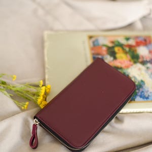 May include: A burgundy leather wallet with a black zipper and a matching pull tab. The wallet is rectangular and closed. A vintage floral print is in the background, along with yellow flowers.