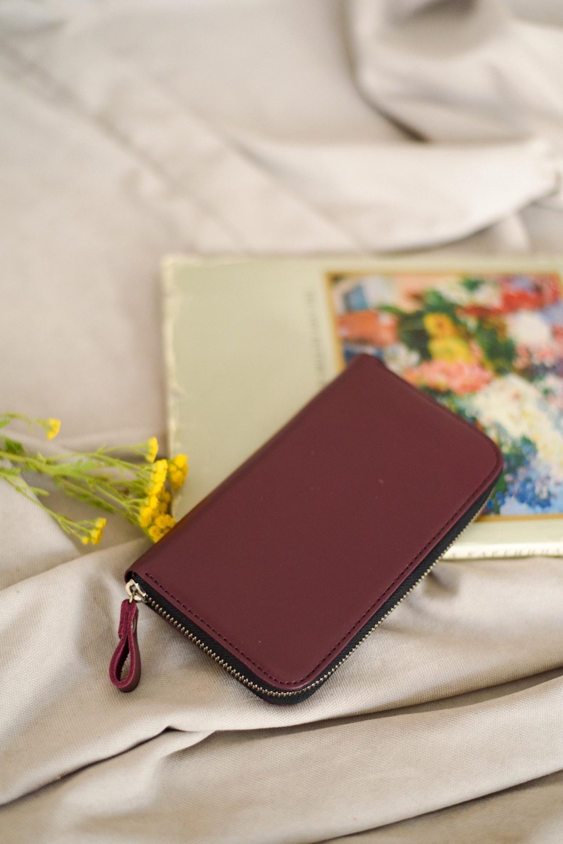 May include: A burgundy leather wallet with a black zipper and a matching pull tab. The wallet is rectangular and closed. A vintage floral print is in the background, along with yellow flowers.
