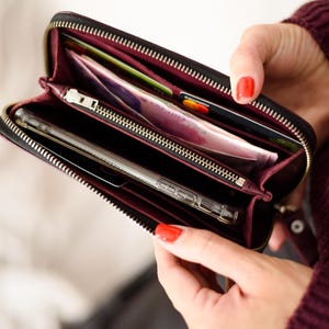 May include: Black zippered wallet with a burgundy interior, holding a smartphone, credit cards, and cash. The wallet is open, revealing multiple compartments. The zipper is gold-colored.