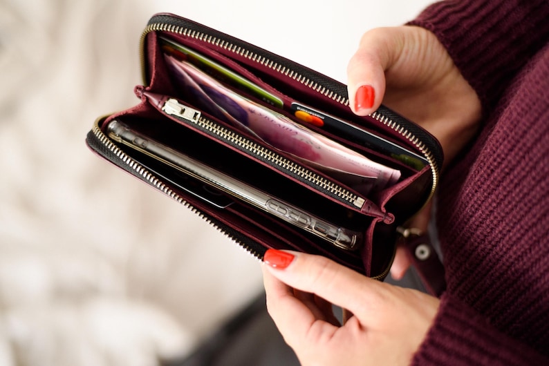 May include: Black zippered wallet with a burgundy interior, holding a smartphone, credit cards, and cash. The wallet is open, revealing multiple compartments. The zipper is gold-colored.