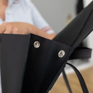 May include: Close-up of a black leather tote bag with silver snap closures. The bag has a simple, modern design with a structured shape and a long strap. The focus is on the bag's details, highlighting the quality of the leather and the hardware.