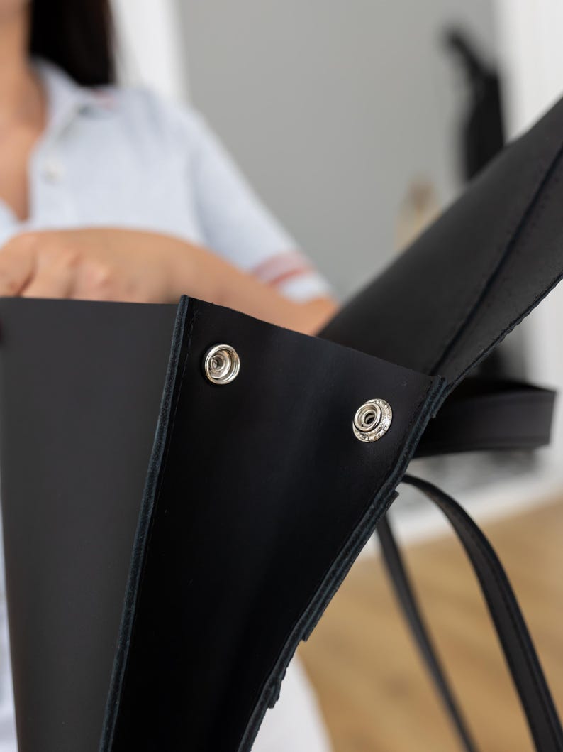 May include: Close-up of a black leather tote bag with silver snap closures. The bag has a simple, modern design with a structured shape and a long strap. The focus is on the bag's details, highlighting the quality of the leather and the hardware.