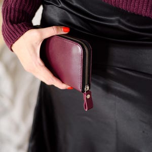 May include: A burgundy leather wallet with a zipper closure and a small wrist strap. The wallet is held against a black satin skirt. The person holding the wallet is wearing a burgundy sweater.